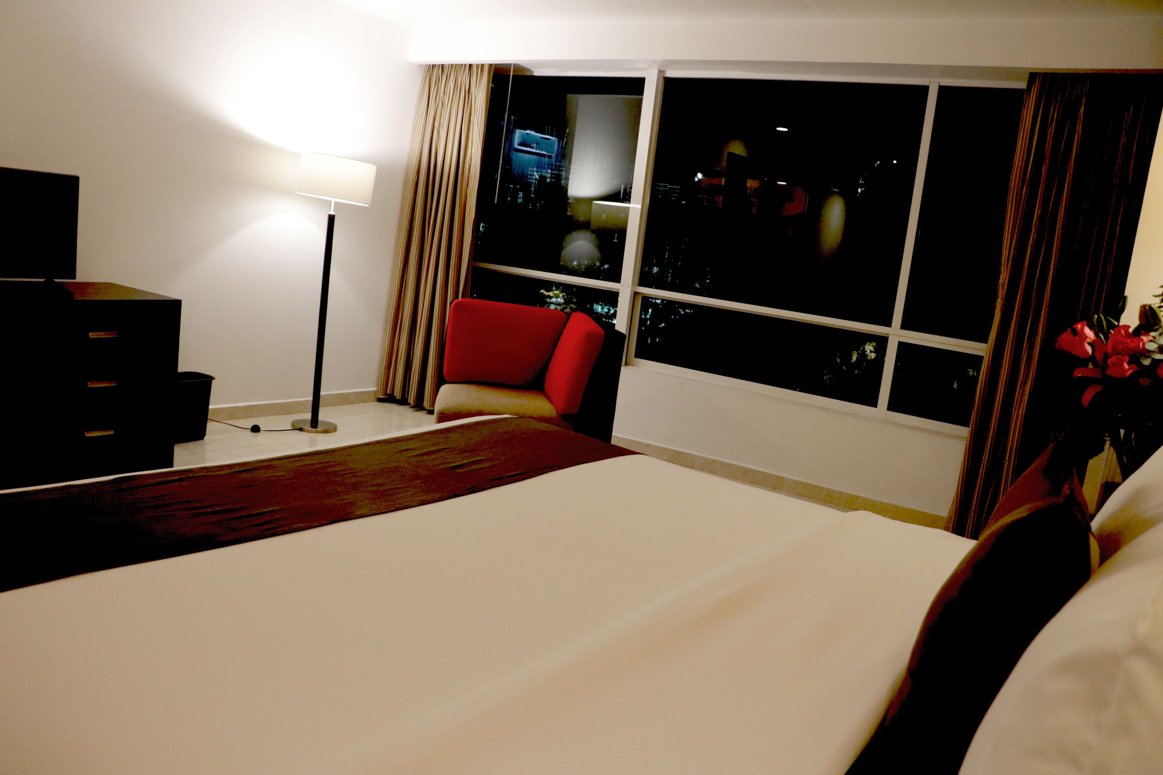 Une chambre d’hôtel avec un grand lit, un fauteuil rouge, un bureau avec une lampe et une vue sur la ville la nuit.