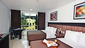 A hotel room with two beds, a desk, and a large window offering a view of buildings and trees.