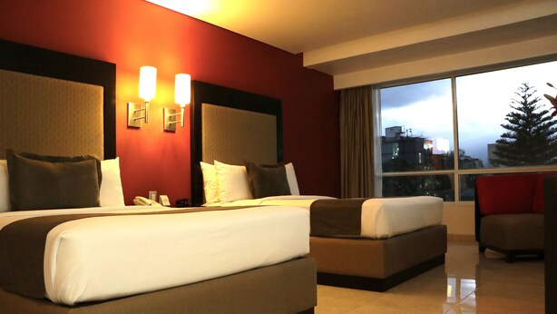 A hotel room with two beds, a sofa, and a chair. There are two wall-mounted lamps and a window with a view of buildings and a tree.
