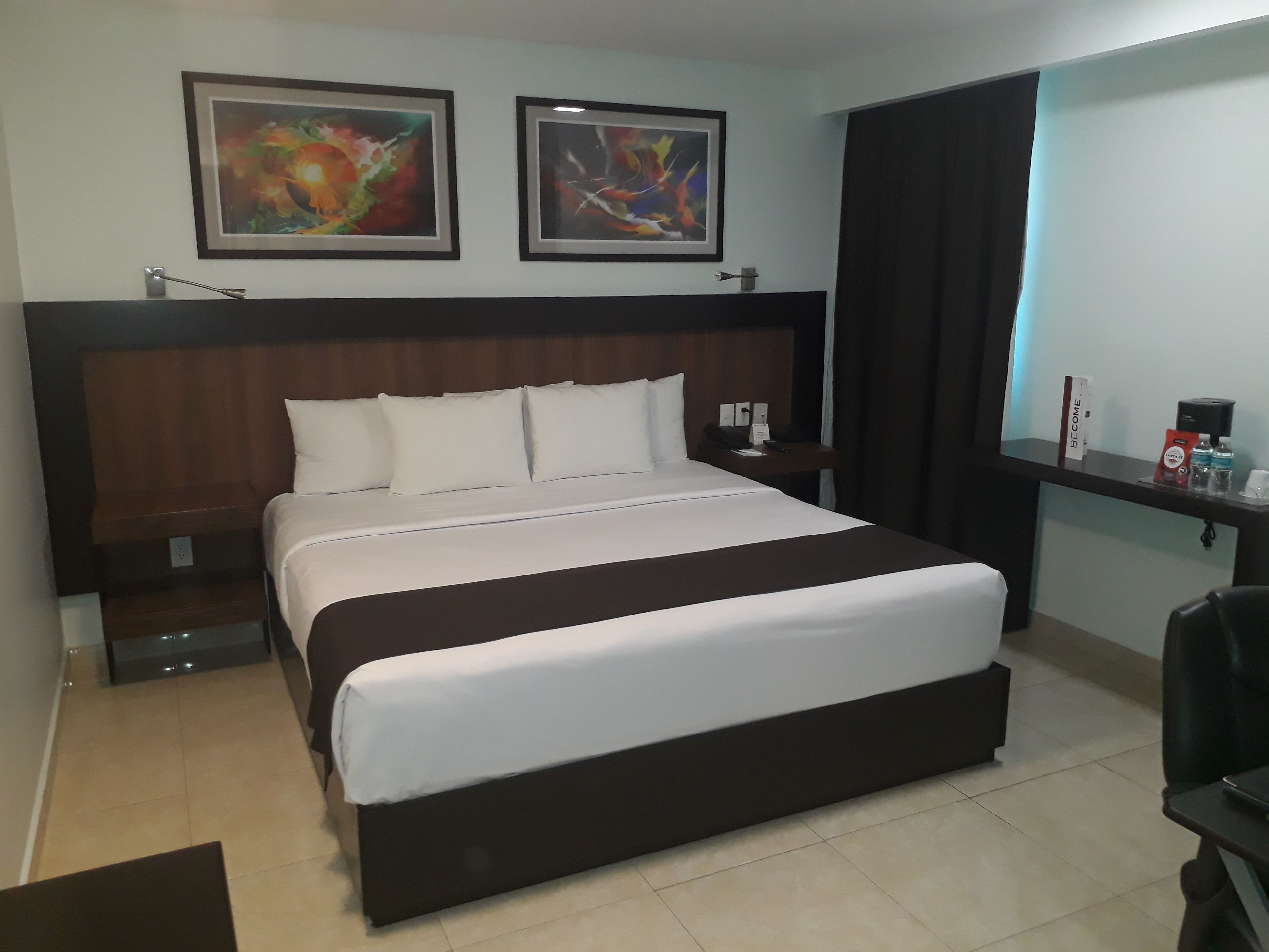 A hotel room with a large bed, a wooden headboard, two abstract paintings on the wall, a nightstand with a lamp, and a desk with a coffee machine.