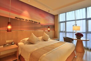 A hotel room with a large bed, a nightstand, a desk with a lamp, and a view of the city through large windows. - Yiwu International Mansion Hotel (Jinhua)