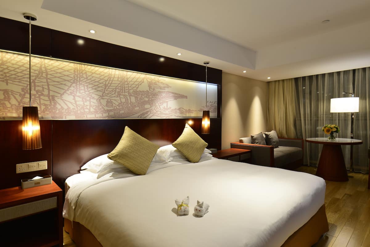 A hotel room with a large bed, two bedside lamps, a nightstand, a small table with a lamp, a chair, and a decorative wall piece.