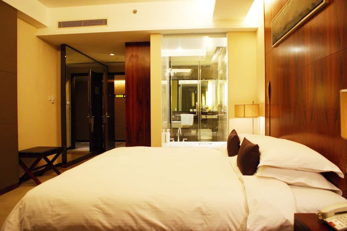 A hotel room with a large bed, a bedside telephone, and a glass door leading to a bathroom.