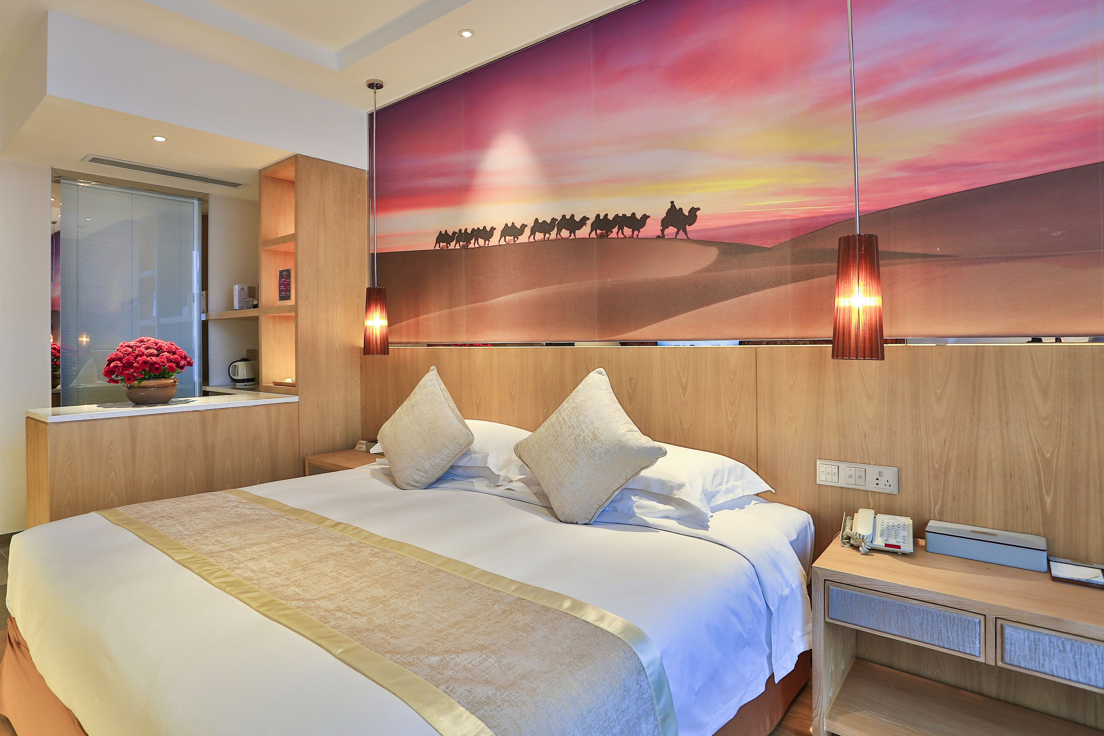 Una habitación de hotel con una cama grande, una mesita de noche con lámpara, un ventanal con vistas y una pared con un cuadro de atardecer.