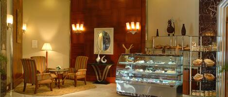 A lobby with a marble floor, a seating area with chairs, a glass display case with pastries, and wall sconces.