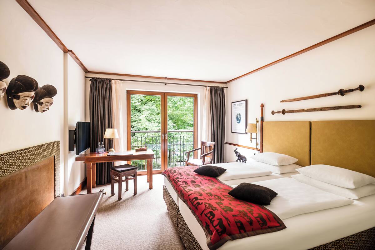 Une chambre d’hôtel comprenant un lit, un bureau, une chaise, un balcon avec des rideaux et une fenêtre donnant sur un espace verdoyant.
