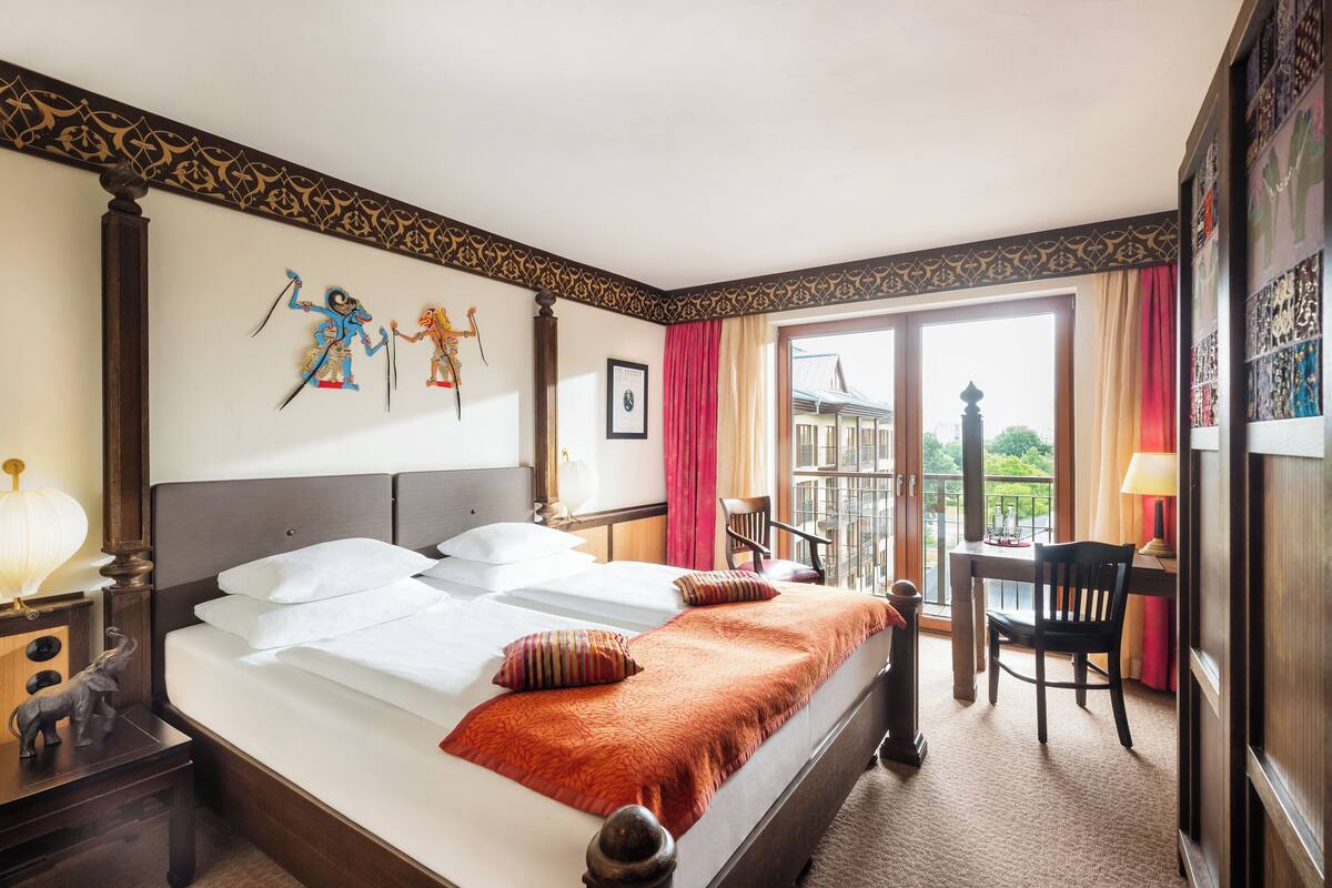 Une chambre d’hôtel avec un grand lit, un bureau et une chaise. Elle dispose d’un balcon avec vue sur les immeubles et les arbres.