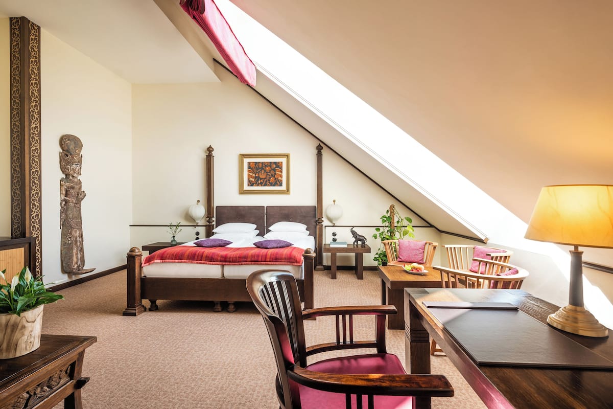 Une chambre avec un plafond en pente, un lit recouvert d’une couverture rouge, un bureau avec une lampe et une chaise.
