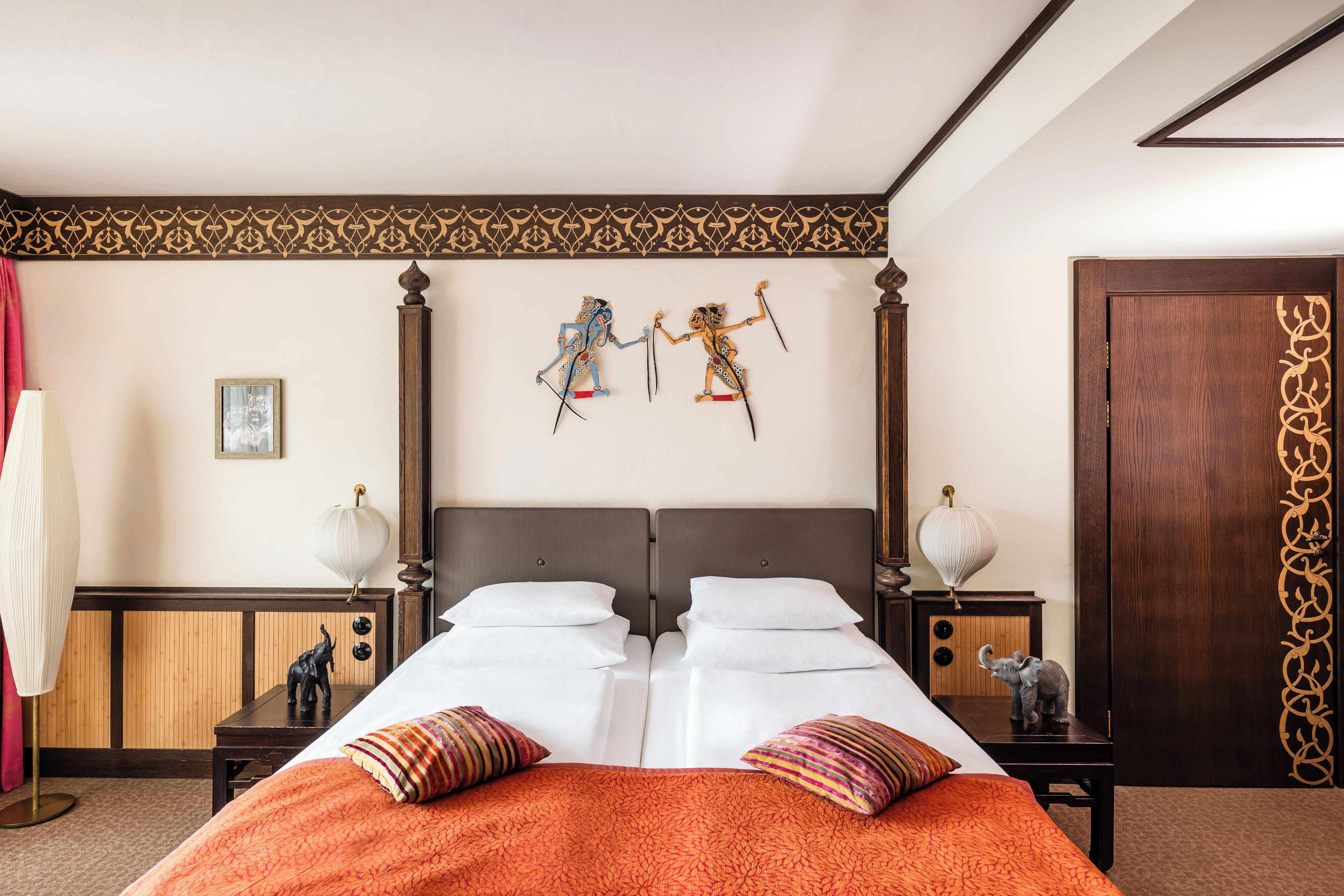Uma cama de casal com colcha laranja, duas mesas de cabeceira com luminárias, um painel de cabeceira de madeira e duas figuras decorativas fixadas na parede.