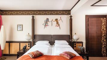 Uma cama de casal com colcha laranja, duas mesas de cabeceira com luminárias, um painel de cabeceira de madeira e duas figuras decorativas fixadas na parede.