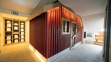 A hallway with a red wall, a wooden door, and a tiled floor. There is a shelf with folded towels and a sign indicating an exit.