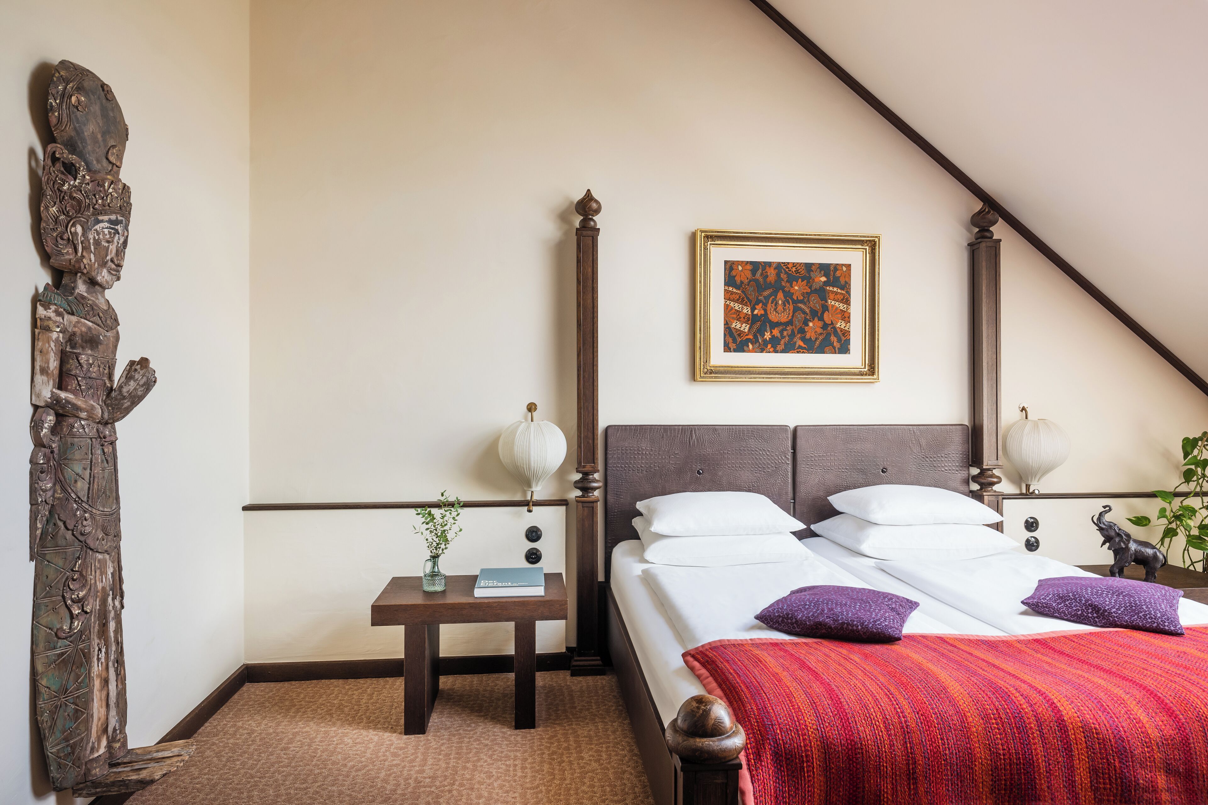 Un dormitorio con una cama, mesitas de noche, una estatua decorativa, una obra de arte enmarcada y una planta.