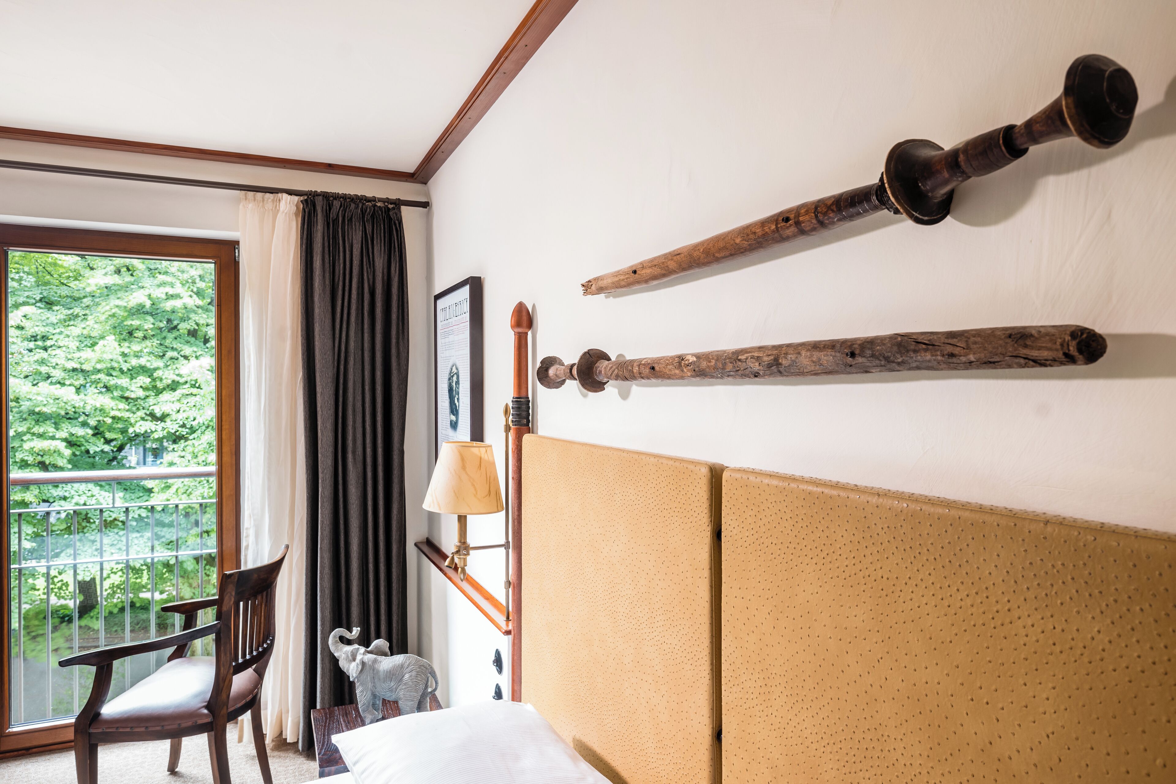 A hotel room with a large window, a wooden chair, a bed with a headboard, and two wooden artifacts on the wall.