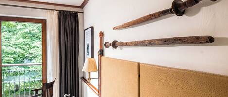 A hotel room with a large window, a wooden chair, a bed with a headboard, and two wooden artifacts on the wall.