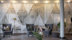 A modern lobby with geometric wall art, grey armchairs, a floor lamp, and a vase with flowers. - Hotel Nickisch (Schuettorf)