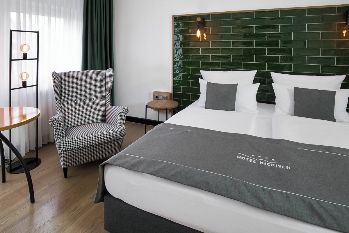 Ein Hotelzimmer mit einem großen Bett, einem karierten Sessel, einem kleinen Tisch und einem dunkelgrünen, mit Fliesen verkleideten Kopfteil.