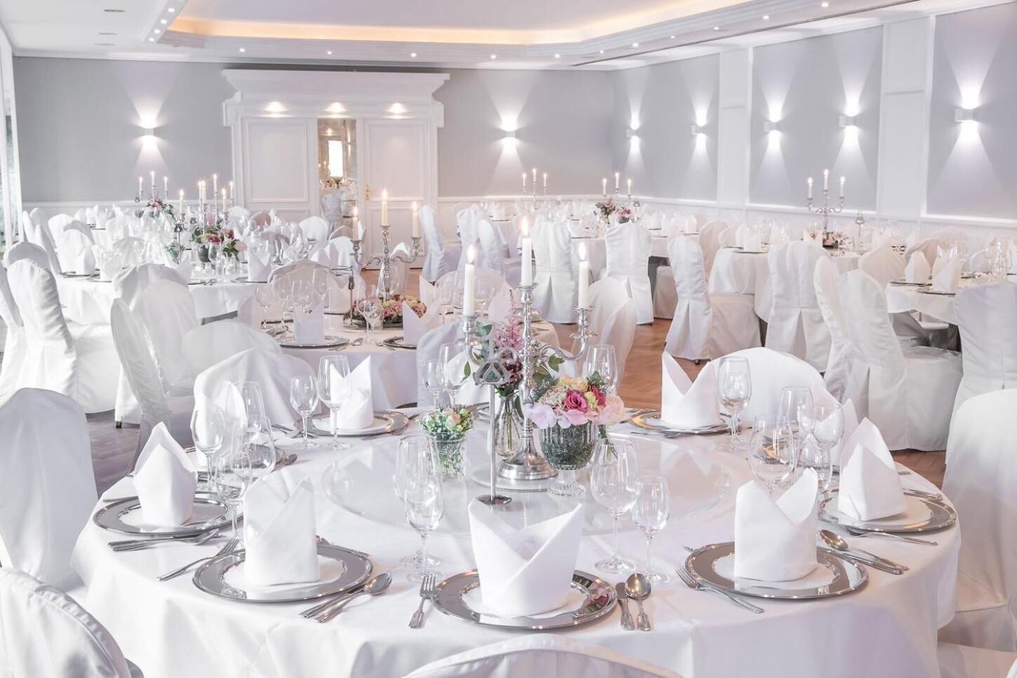 Ein Festsaal mit runden Tischen, gedeckt für eine festliche Veranstaltung, mit weißen Tischdecken, Besteck und floralen Tischarrangements.