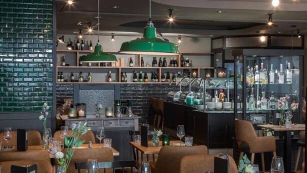A restaurant interior with wooden tables, green pendant lighting, and shelves stocked with bottles. - Hotel Nickisch (Schuettorf)