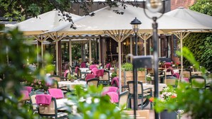 An outdoor café with tables and chairs, covered by umbrellas, and surrounded by greenery. - Hotel Nickisch (Schuettorf)