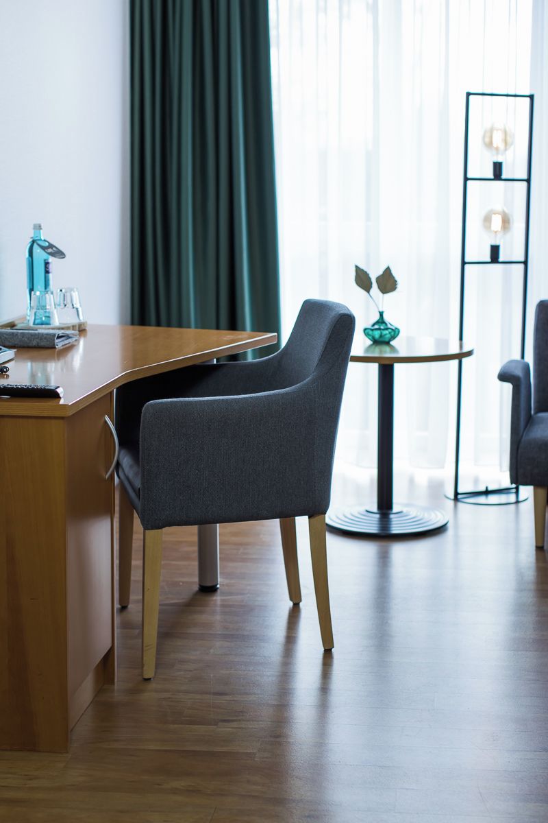 Una habitación de hotel moderna con un escritorio de madera, una butaca tapizada en gris, una pequeña mesa redonda con un jarrón y una lámpara de pie negra alta.