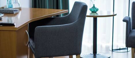 Una habitación de hotel moderna con un escritorio de madera, una butaca tapizada en gris, una pequeña mesa redonda con un jarrón y una lámpara de pie negra alta.