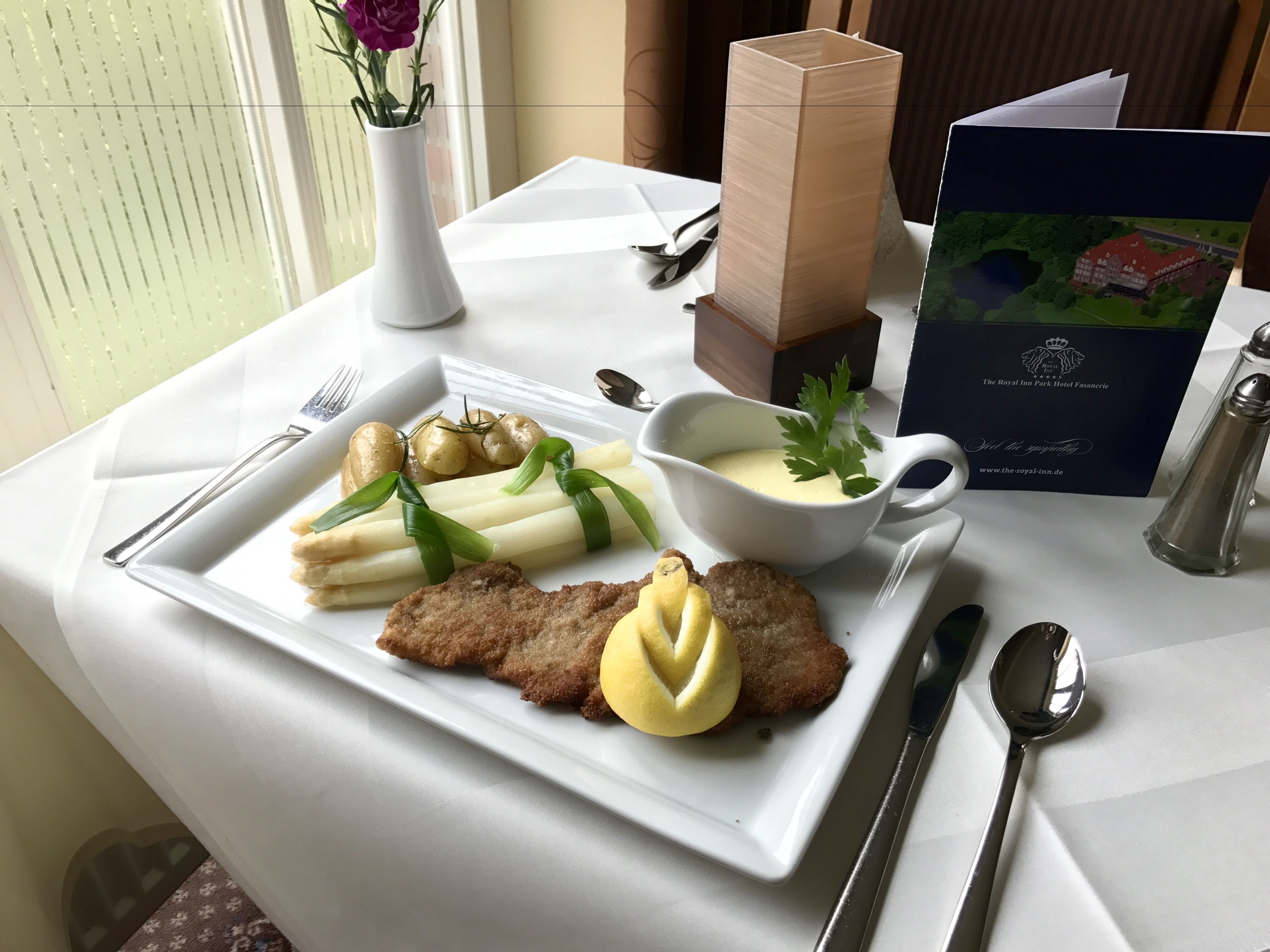 Ein Esstisch mit einem weißen Teller, der eine Panade, Kartoffeln und Spargel zeigt, dazu eine kleine Schüssel Soße und ein Zweig Petersilie.