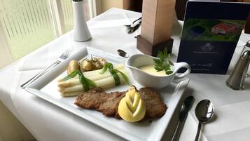 A dining table with a white plate of food, including a breaded cutlet, boiled potatoes, and asparagus, accompanied by a small bowl of sauce and a sprig of parsley.