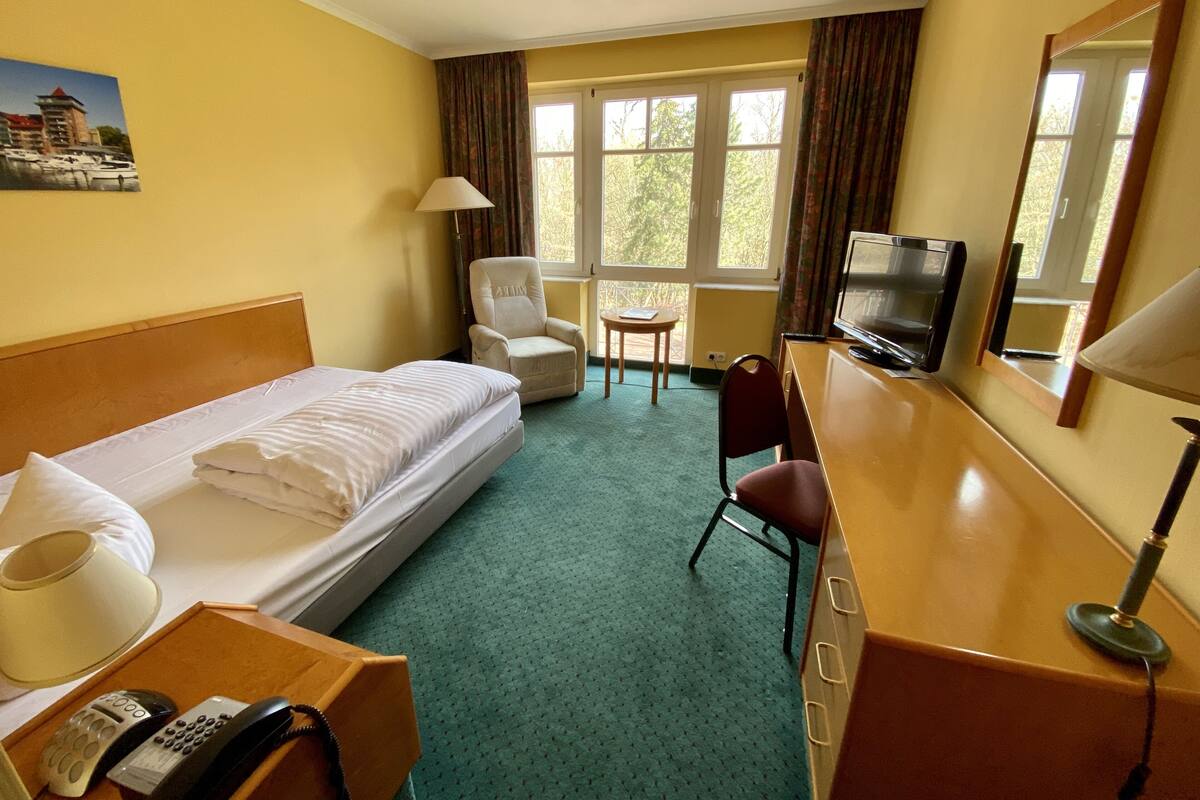 A hotel room with a bed, a desk with a TV, a chair, a small table, and a lamp.