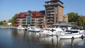 A waterfront hotel with multiple balconies, a red-roofed tower, and boats docked in the water. - Park Hotel Fasanerie Neustrelitz (Neustrelitz)