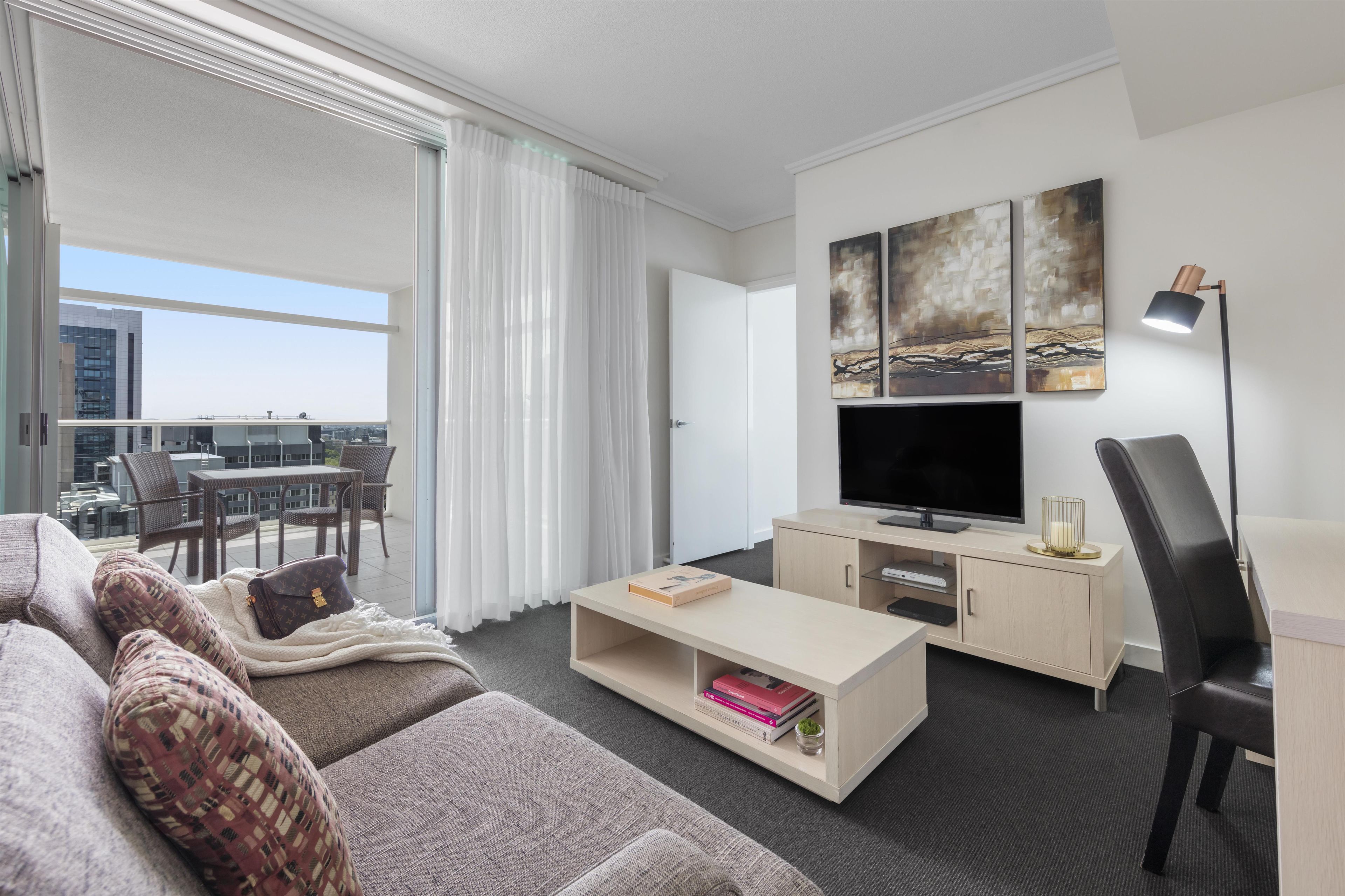 A modern hotel room with a sofa, a TV, a coffee table, and a balcony with a dining area.