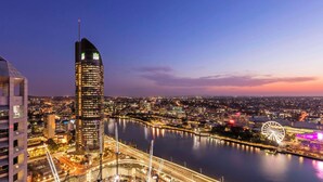 A cityscape at dusk with a prominent skyscraper, a river, and a Ferris wheel. - Oaks Brisbane Casino Tower Suites (Brisbane)