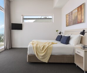 A modern hotel room with a large bed, a flat-screen TV mounted on the wall, a window with a view of a bridge, and a painting above the bed.
