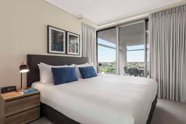 A hotel room with a large bed, a nightstand with a lamp, a digital clock, and a view of the city through a sliding glass door. - Oaks Brisbane Casino Tower Suites (Brisbane)