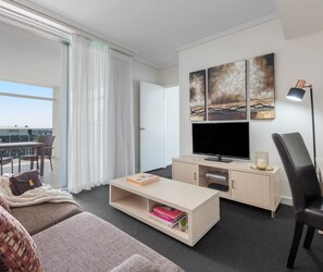 A modern hotel room with a large flat-screen TV, a sofa, a coffee table, and a desk area.