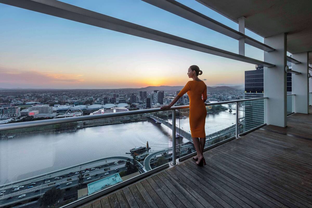 Une femme se tient sur un balcon, d’où elle a une vue sur un paysage urbain avec une rivière et des ponts, au moment du coucher du soleil.