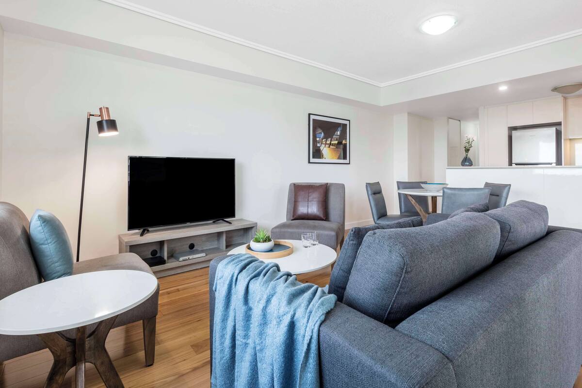 Un salon moderne avec un canapé gris, un téléviseur à écran plat, une table basse et un coin repas avec des chaises.