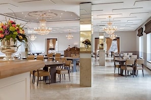 A spacious hotel dining area with multiple tables and chairs, chandeliers, and a large mirror reflecting the space.