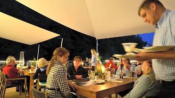 An outdoor dining area with tables and chairs, a waiter serving food, and a person in the foreground.