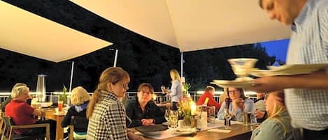 Un espace extérieur aménagé pour les repas, avec des tables et des chaises, un serveur qui apporte les plats, et une personne au premier plan.