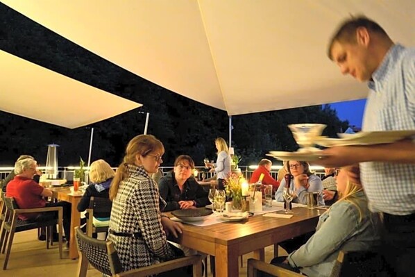 An outdoor dining area with tables and chairs, a waiter serving food, and a person in the foreground.