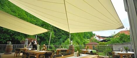 Un espace extérieur aménagé en terrasse, avec des tables et des chaises en bois, un grand parasol pour faire de l’ombre, et une vue sur un paysage verdoyant.