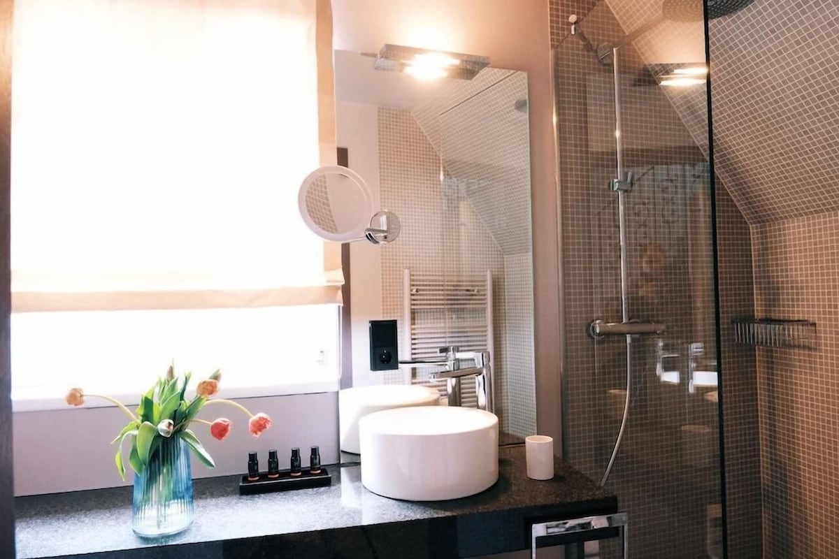 A modern bathroom with a glass-enclosed shower, a round mirror, and a vase with flowers on the counter.
