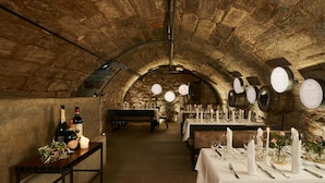 Un comedor subterráneo con paredes de piedra, mesas redondas dispuestas con manteles blancos, velas y botellas de vino.