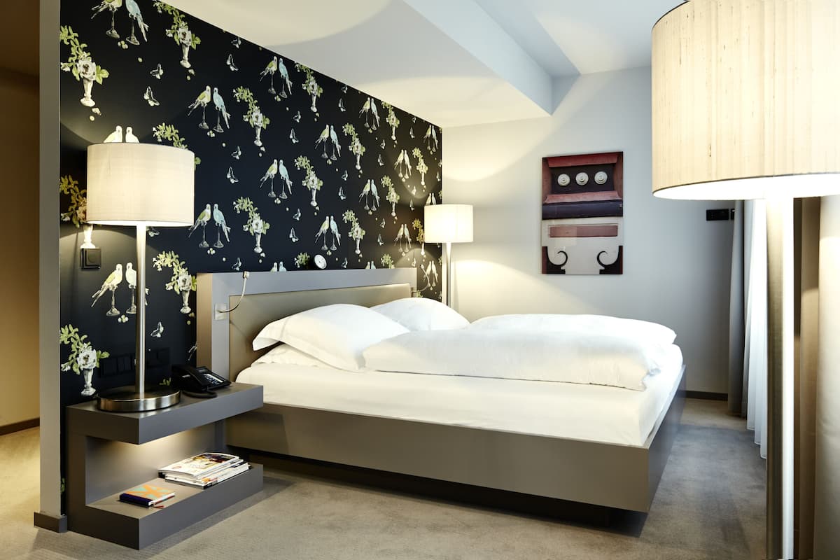 A modern hotel room with a large bed, two bedside lamps, a nightstand with a phone, and a wall with a bird pattern.