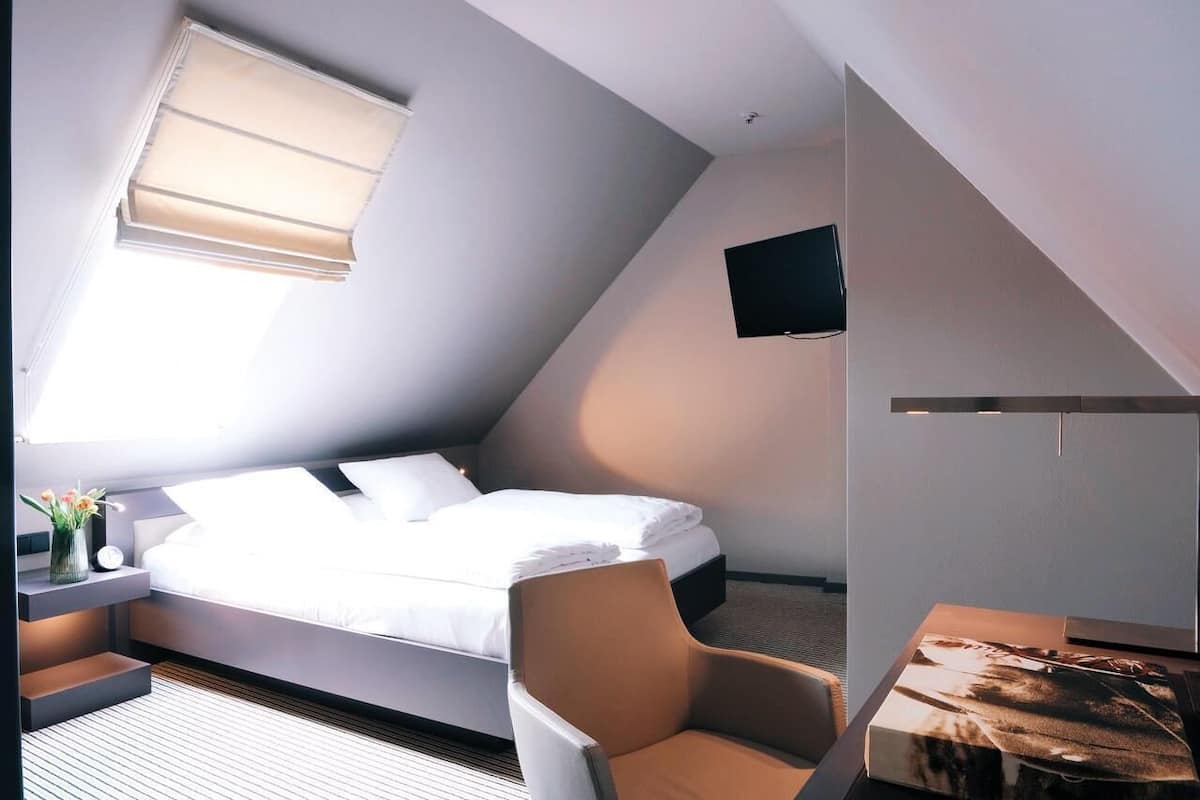 A modern, compact room with a bed, a chair, a small table, and a TV mounted on the wall.