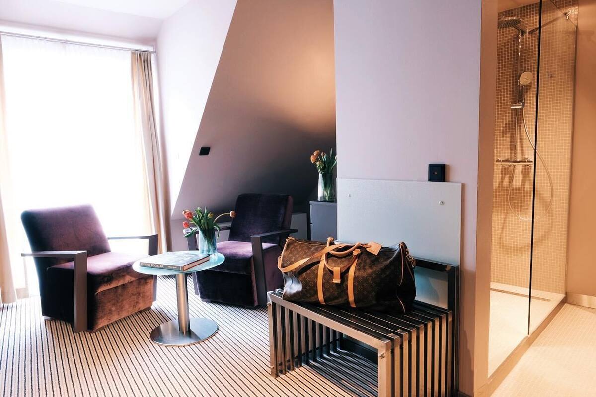 A modern hotel room with a large window, two armchairs, a small round table, a minibar, and a shower area.