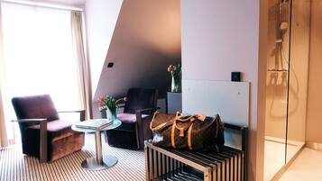 A modern hotel room with a large window, two armchairs, a small round table, a minibar, and a shower area.