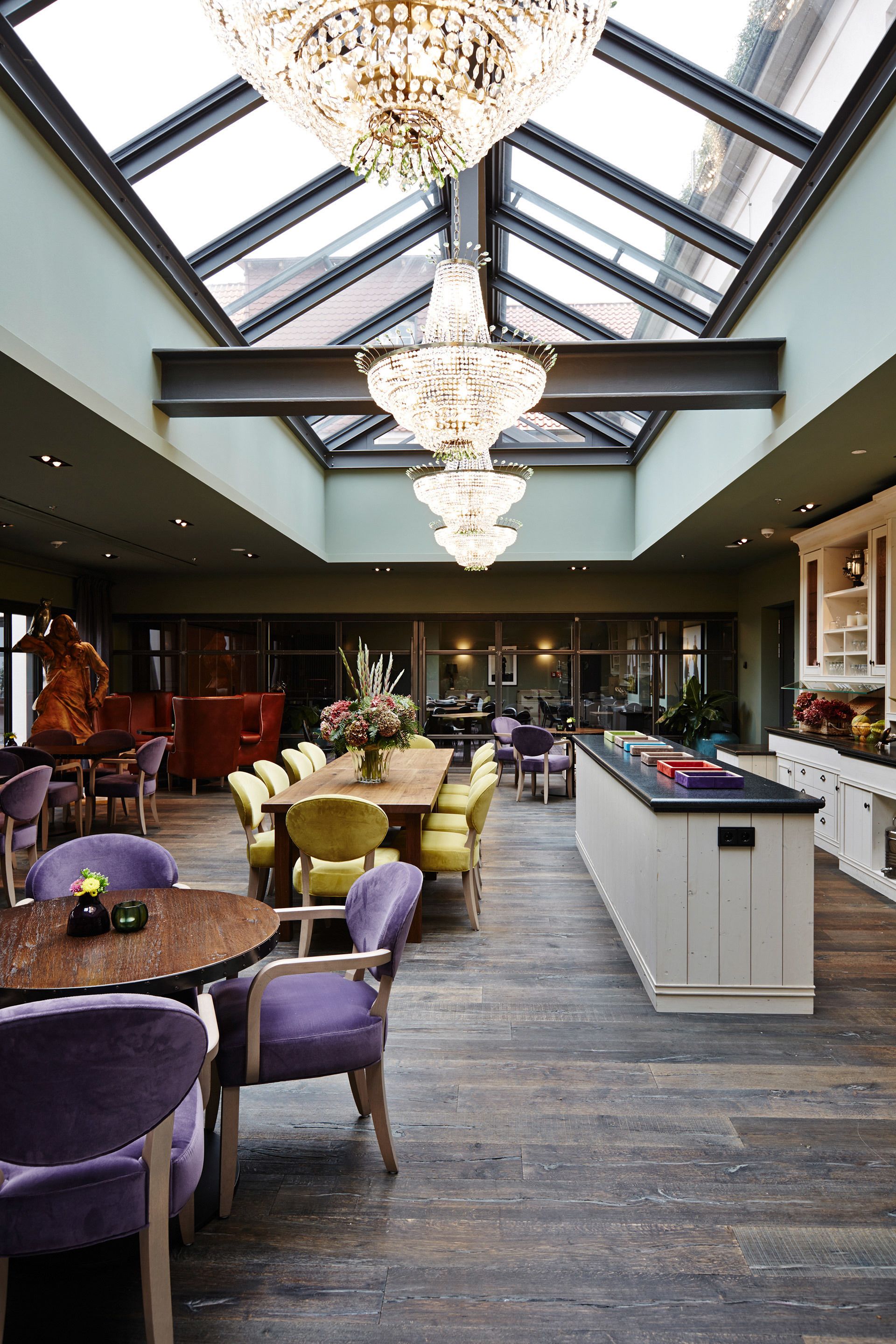 A spacious interior with a high ceiling, a central dining area with yellow chairs, a chandelier, and a kitchen with a large island.