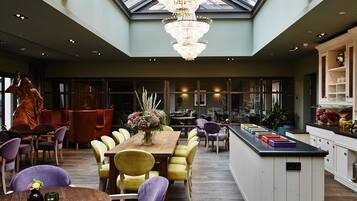 A spacious interior with a high ceiling, a central dining area with yellow chairs, a chandelier, and a kitchen with a large island.