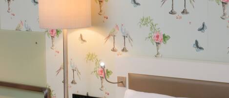 A hotel room with a bed, a bedside table with a lamp, a clock, and a wall with floral and bird wallpaper.